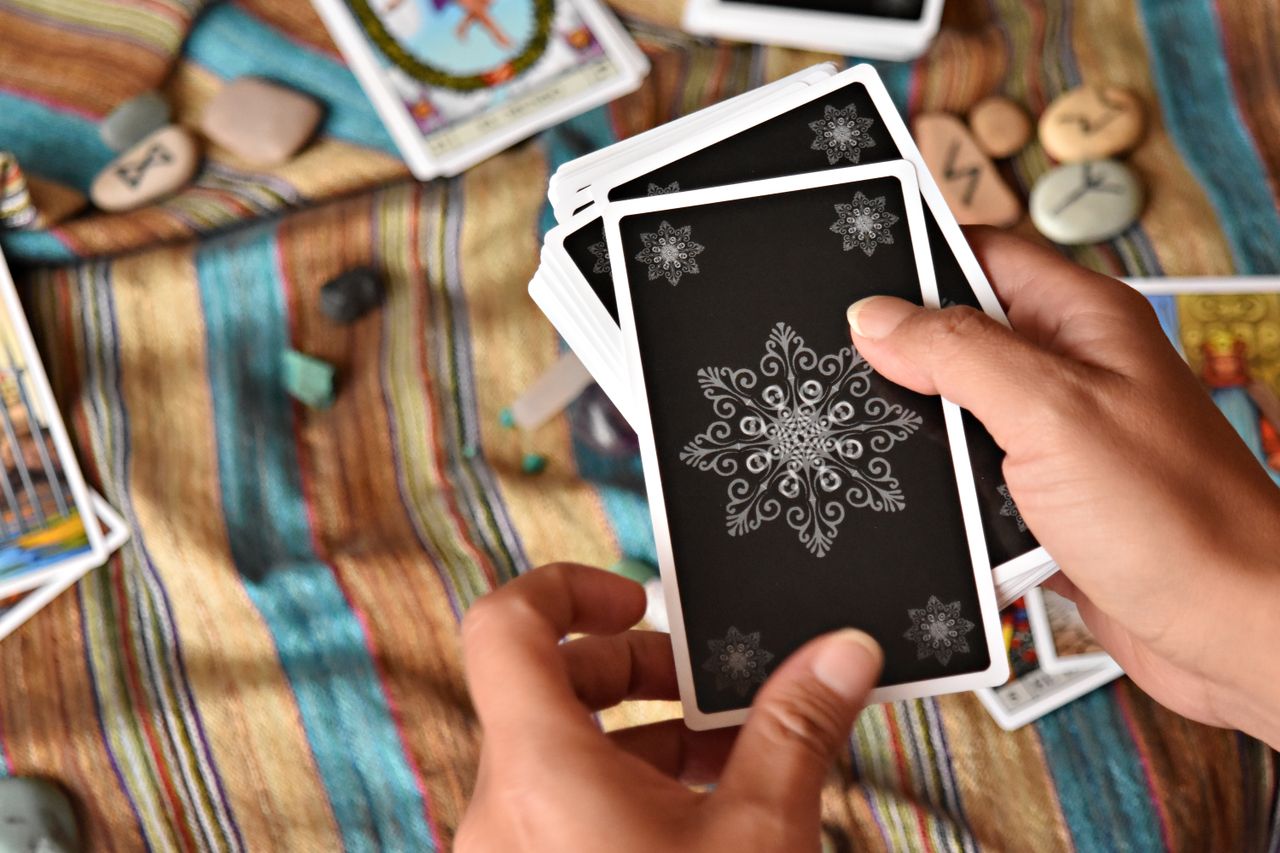 Hands holding tarot cards on a colorful textured cloth, preparing for a reading with stones and runes.